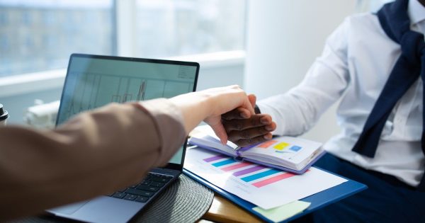 Two professionals shaking hands across a desk covered with charts and reports beside a laptop