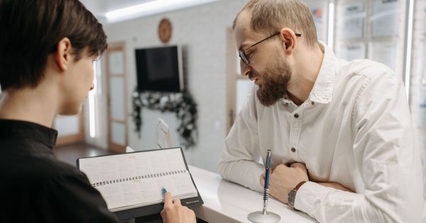 A clinic receptionist points to a date in an appointment calendar while a patient looks on at the front desk